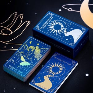 Pode incluir: Uma caixa azul com letras douradas e ilustra&ccedil;&otilde;es de um sol e uma lua. A caixa cont&eacute;m um baralho de cartas de tar&ocirc; com um design azul e dourado. O texto na caixa diz "The Golden Path Tarot: An Acolyte Deck and Guidebook".