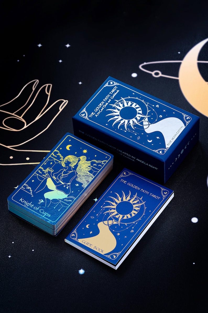 Pode incluir: Uma caixa azul com letras douradas e ilustra&ccedil;&otilde;es de um sol e uma lua. A caixa cont&eacute;m um baralho de cartas de tar&ocirc; com um design azul e dourado. O texto na caixa diz "The Golden Path Tarot: An Acolyte Deck and Guidebook".