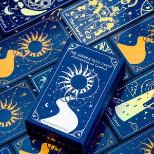 Pode incluir: Uma caixa azul com letras douradas que dizem "The Golden Path Tarot A 78-Card Deck and Guidebook Blue Moon Edition". A caixa &eacute; cercada por cartas de tar&ocirc; com fundos azuis e desenhos dourados.