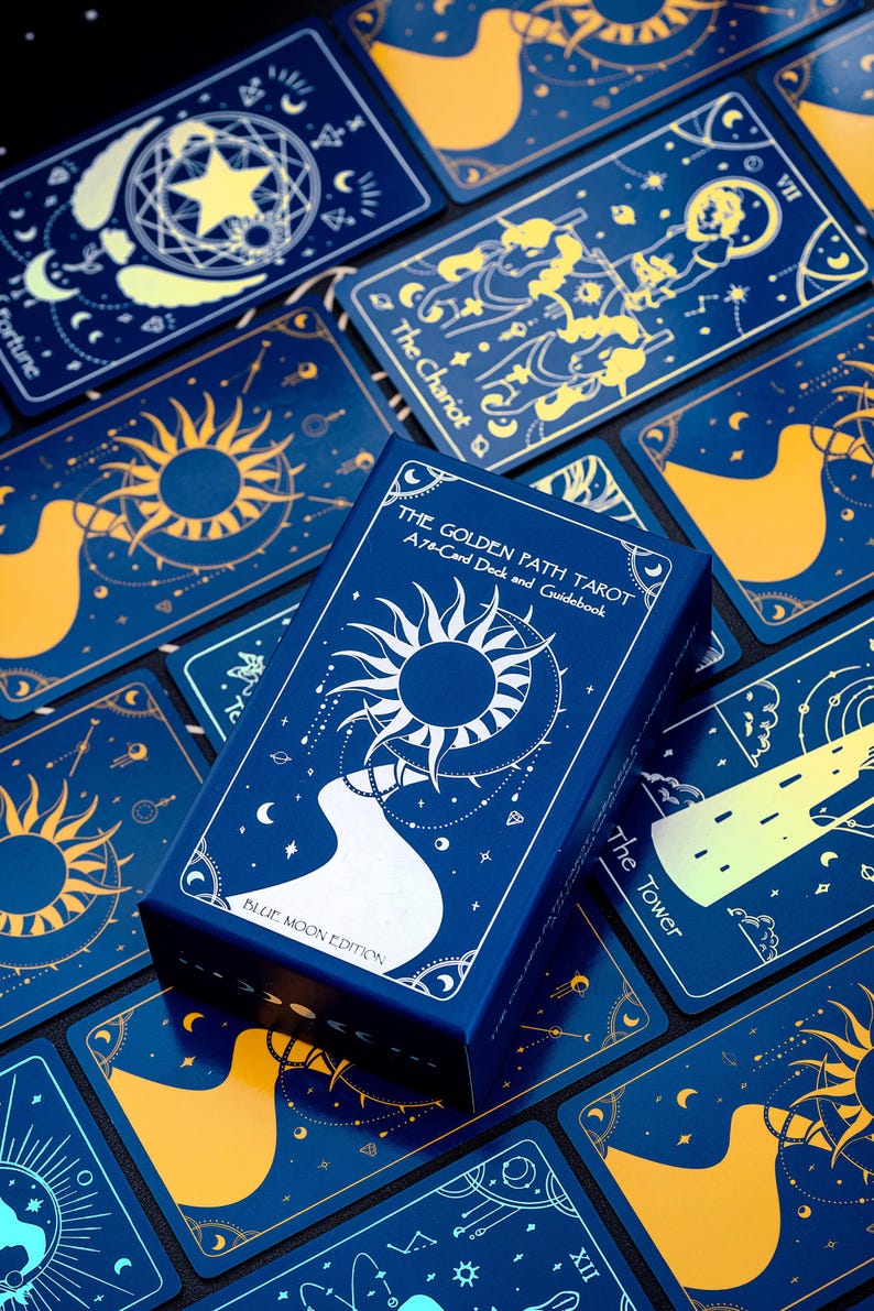 Pode incluir: Uma caixa azul com letras douradas que dizem "The Golden Path Tarot A 78-Card Deck and Guidebook Blue Moon Edition". A caixa &eacute; cercada por cartas de tar&ocirc; com fundos azuis e desenhos dourados.
