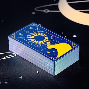 Pode incluir: Um baralho de cartas de tarot com fundo azul e detalhes dourados. A carta superior apresenta um desenho de sol e lua.