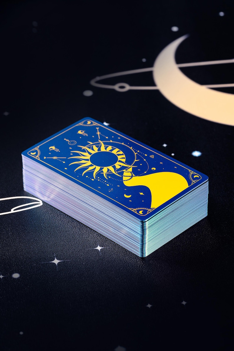 Pode incluir: Um baralho de cartas de tarot com fundo azul e detalhes dourados. A carta superior apresenta um desenho de sol e lua.