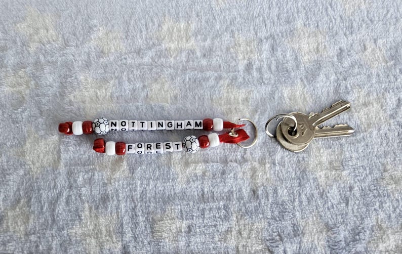 Puede incluir: Un llavero de cuentas rojas y blancas con las palabras "NOTTINGHAM FOREST" y un peque&ntilde;o charm de bal&oacute;n de f&uacute;tbol. El llavero est&aacute; unido a un llavero plateado con dos llaves.