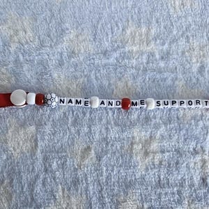 Puede incluir: Un clip para chupete blanco con detalles rojos y cuentas blancas. El clip tiene el texto "NAME AND ME SUPPORT" impreso en &eacute;l.