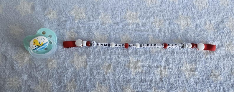 Puede incluir: Un clip para chupete blanco con detalles rojos y cuentas blancas. El clip tiene el texto "NAME AND ME SUPPORT" impreso en &eacute;l.