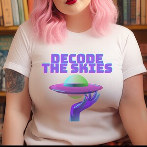 Puede incluir: Camiseta blanca con un gráfico de una mano sosteniendo un platillo volante morado y verde. El texto "DECODE THE SKIES" está impreso en morado y azul.