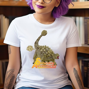 Może przedstawiać: Biała koszulka z kreskówkową ilustracją zielonego dinozaura Ankylozaura z chmurą dymu za nim. Tekst "Saturday Adventures" jest nadrukowany pod dinozaurem.