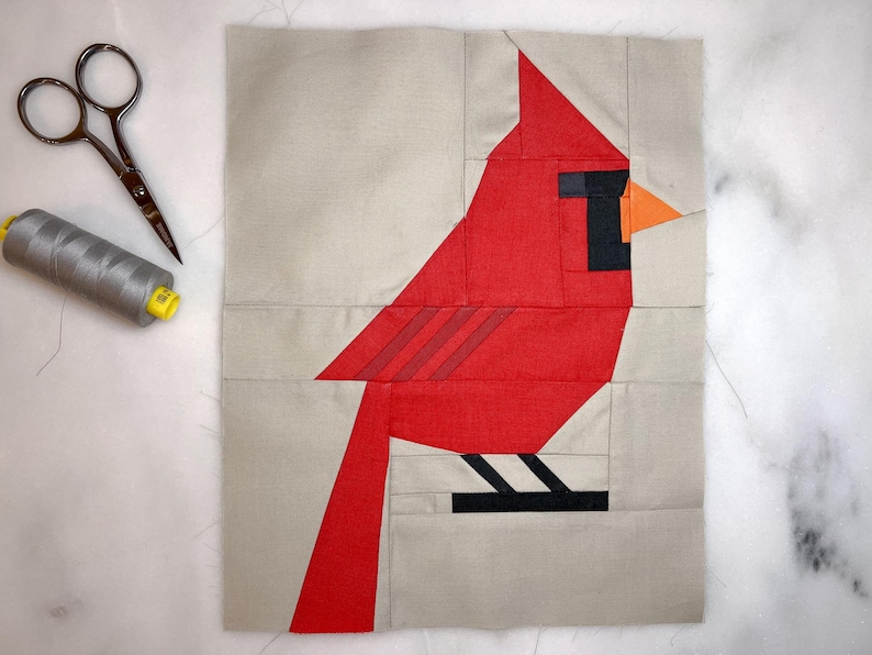 May include: A red cardinal bird applique made from fabric pieces. The bird is on a beige background. The bird has a black eye and beak, and an orange beak.