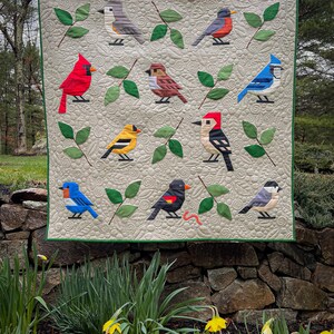 May include: A quilt featuring a variety of colorful birds, including a cardinal, blue jay, woodpecker, and goldfinch, against a cream background with green leaves.