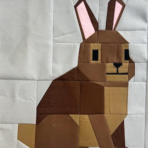 May include: A geometric patchwork design featuring a brown rabbit with pink-lined ears. The rabbit is set against a light gray background, with the design constructed from various fabric squares. The rabbit's eyes and nose are black.