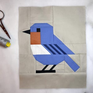 May include: A bluebird quilt block with a white chest and black stripes. The bird is on a beige background.
