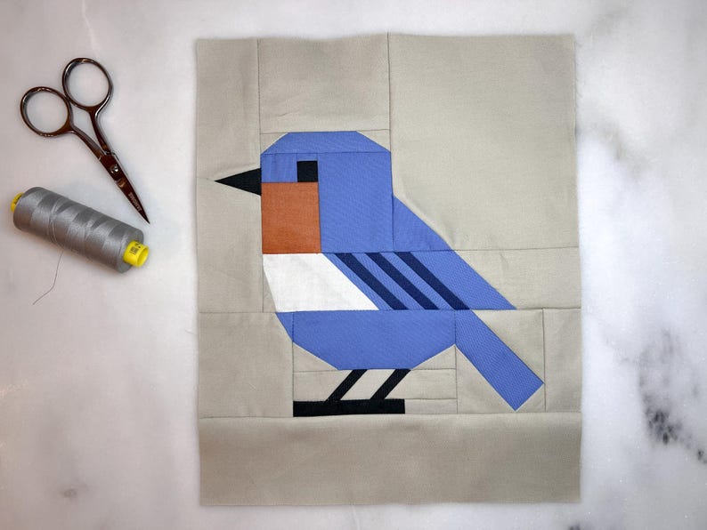 May include: A bluebird quilt block with a white chest and black stripes. The bird is on a beige background.