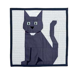 May include: A square quilt featuring a geometric design of a dark blue cat with a white collar, set against a light gray background with a subtle pattern. The quilt is framed by a dark blue border.