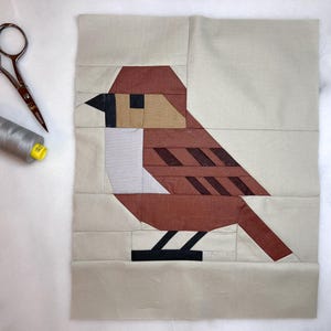 May include: A brown and white patchwork bird with black feet and a beige background. The bird is made of fabric and is a quilt block.
