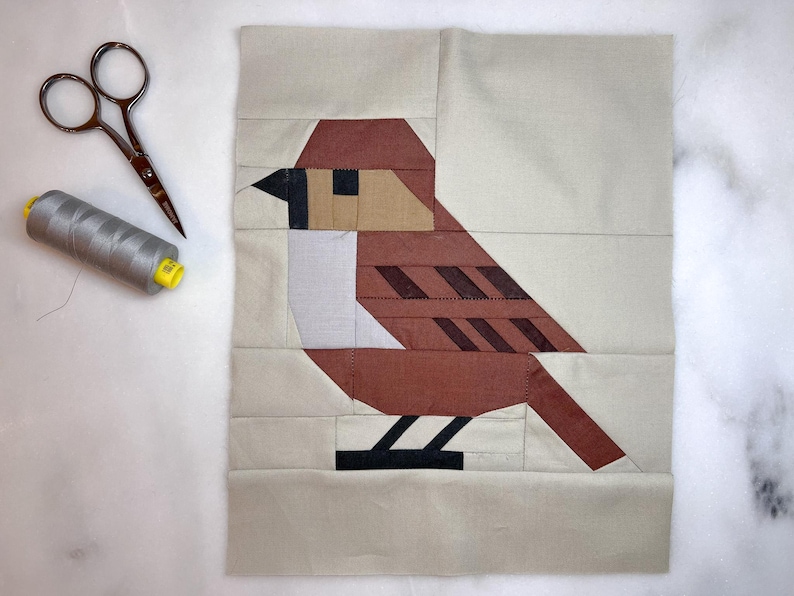 May include: A brown and white patchwork bird with black feet and a beige background. The bird is made of fabric and is a quilt block.