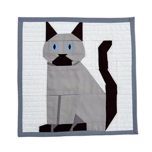 May include: A Siamese cat quilt with a gray body, black ears and tail, and blue eyes. The cat is sitting upright on a white background with a gray border. The quilt is made of fabric pieces.
