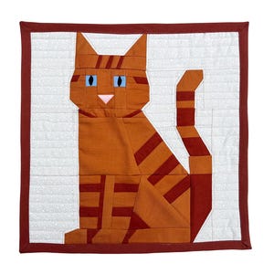 Puede incluir: Un edredón hecho a mano con un gato naranja geométrico con rayas naranja oscuro, ojos azules y nariz rosa. El gato está centrado en un fondo blanco con un sutil patrón de puntos, enmarcado por un borde rojo oscuro.