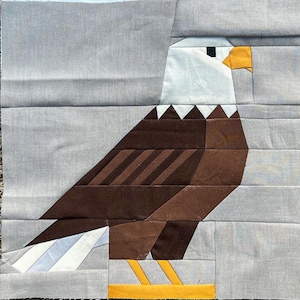 Op de afbeelding: Een stoffen patchworkblok met een gestileerd adelaarsontwerp. De adelaar heeft een witte kop, een bruin lichaam en een gele snavel en poten. De achtergrond is een lichtgrijze stof.