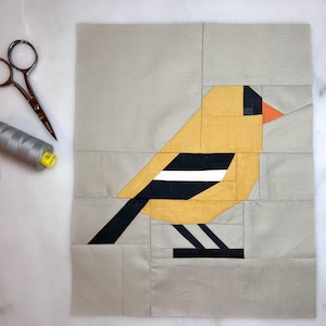 May include: A patchwork quilt block featuring a geometric design of a yellow bird with black and white wings and an orange beak. The bird is set against a light gray background.