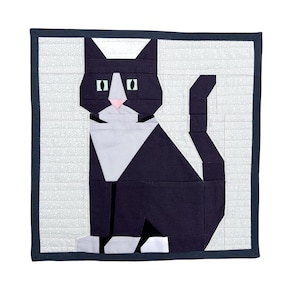 Könnte beinhalten: Eine gesteppte Wandbehänge mit einem geometrischen Design einer schwarz-weißen Katze. Die Katze hat eine rosa Nase und grüne Augen. Der Hintergrund ist weiß mit einem dezenten Muster, umrahmt von einem dunkelblauen Rand.
