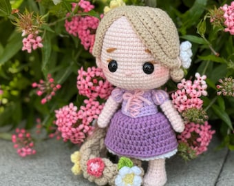 May include: A crocheted doll with long blonde hair, wearing a purple dress and a flower crown. The doll is surrounded by pink flowers.
