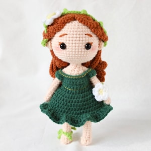May include: A crocheted doll with brown hair, a green dress, and a white flower. The doll is wearing a green leaf crown.