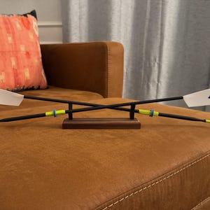 May include: A decorative rowing oar model. The oars are black with white and blue tips, yellow and blue accents, and a wooden base. The model is displayed on a brown surface.