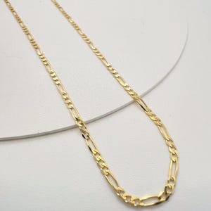 May include: A gold-colored figaro chain necklace featuring a repeating pattern of three small circular links followed by one elongated oval link. The metal chain has a polished, reflective finish and is displayed in a simple, elegant loop.