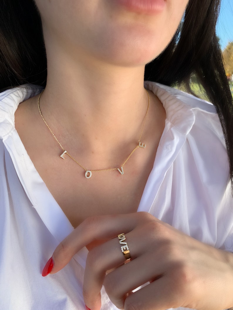 May include: A gold necklace with the word "LOVE" spelled out in diamonds. The necklace is worn by a person wearing a white shirt. The person is also wearing a gold ring with the word "LOVE" spelled out in diamonds.