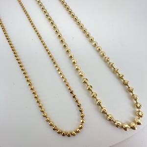 May include: A selection of gold-coloured necklaces with varying bead designs are presented against a white backdrop. The necklaces showcase different bead sizes and shapes, offering a contrast in texture and style. The chains appear delicate and are likely made of metal.