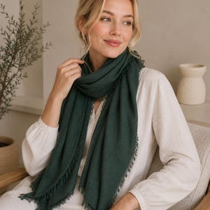 Puede incluir: Una bufanda de color verde bosque oscuro con un delicado detalle de bordes deshilachados. La bufanda está colocada alrededor del cuello y los hombros, mostrando un acabado de tela texturizada. El color verde intenso contrasta con la blusa clara.