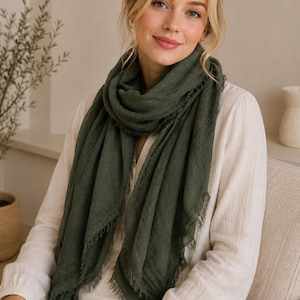 May include: A dark olive green scarf with a textured, lightweight fabric and frayed edges. The scarf is draped loosely around the neck, featuring a rectangular shape with visible woven threads and a soft, slightly wrinkled appearance throughout.