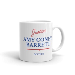 May include: White ceramic coffee mug with a blue and red design. The mug features the text "Justice Amy Coney Barrett SCOTUS".