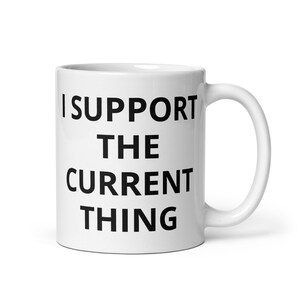Könnte beinhalten: Weiße Keramik-Kaffeetasse mit schwarzem Text, der "Ich unterstütze das aktuelle Ding" lautet.