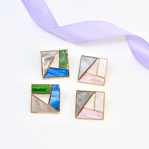 May include: Four square-shaped earrings with gold-colored frames. Each earring features a geometric design with sections of white, gray, green, blue, and pink. A lavender ribbon is in the background.