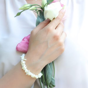 May include: A white shell bracelet with irregular shaped pieces of shell. The bracelet is on a person's wrist, and the person is holding a bouquet of white and pink flowers.