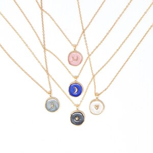 May include: Five gold necklaces with different pendants. Each pendant is a round, white, enamel-like disc with a different gold design: a crescent moon, a star, a lightning bolt, a heart, and a crescent moon.