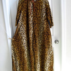 May include: A long-sleeved, floor-length dress featuring a brown, tan, and black leopard print pattern. The garment has a round neckline with a small front slit and a tie closure at the collar, hanging from a white plastic hanger.