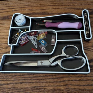 May include: A black and white sewing organiser shaped like a sewing machine, filled with sewing tools. Includes scissors, measuring tape, pins, seam rippers, and various clips. The organiser sits on a wooden surface.