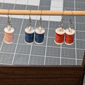 Puede incluir: Seis pares de pendientes en un expositor de madera. Cada par presenta un pequeño carrete de hilo, con colores que incluyen rosa, azul y rojo.