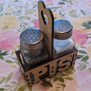 May include: Wooden salt and pepper shaker holder with a heart-shaped handle. The holder has the word "P.S." cut out on the front.