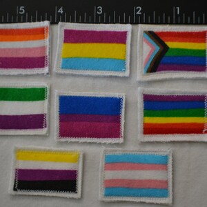May include: Nine different pride flag patches, each with a different color scheme and design. The patches are sewn onto a white fabric background.
