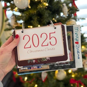 May include: A Christmas card holder with the year 2025 in red script. The holder is open, revealing several cards with family photos. The words "Christmas Cards" are below the year. The holder is held in front of a decorated Christmas tree.