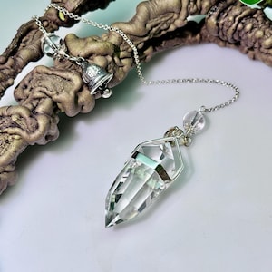 May include: A clear, faceted crystal pendulum pendant suspended from a silver-toned metal chain. The chain features a small, clear spherical bead and a decorative silver-toned bell charm. The crystal is held by a silver-toned metal cap.