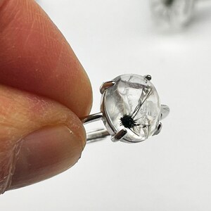 K&ouml;nnte beinhalten: Ein Silberring mit einem klaren, ovalen Edelstein mit schwarzen Einschl&uuml;ssen. Der Ring hat ein schlichtes Band und vier Krappen, die den Stein halten. Der Edelstein zeigt ein unverwechselbares, nat&uuml;rliches Muster.