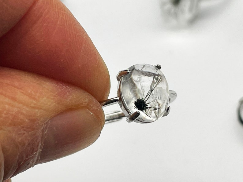 K&ouml;nnte beinhalten: Ein Silberring mit einem klaren, ovalen Edelstein mit schwarzen Einschl&uuml;ssen. Der Ring hat ein schlichtes Band und vier Krappen, die den Stein halten. Der Edelstein zeigt ein unverwechselbares, nat&uuml;rliches Muster.