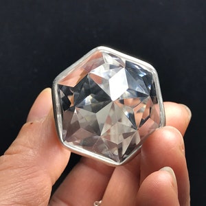 May include: A clear crystal gemstone with a faceted, geometric shape, set in a silver-toned metal frame. The crystal is held in a hand.