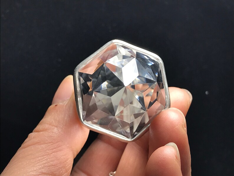 May include: A clear crystal gemstone with a faceted, geometric shape, set in a silver-toned metal frame. The crystal is held in a hand.