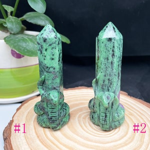 May include: Two green and black serpentine carved crystal points, each with a snake design. The points are on a wooden surface with the numbers #1 and #2 written on the surface.
