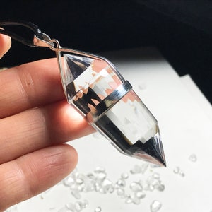Może przedstawiać: Wisiorek z przezroczystego kryształu kwarcowego z srebrną oprawą, wiszący na czarnym sznurku. Kryształ jest fasetowany i ma ostry czubek.